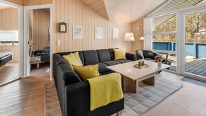 Een moderne woonkamer met een zwarte bank, gele kussens, een houten salontafel en een groot raam met uitzicht op de omgeving.