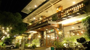 A hotel with a stone facade, wooden balcony, and outdoor seating area with plants.