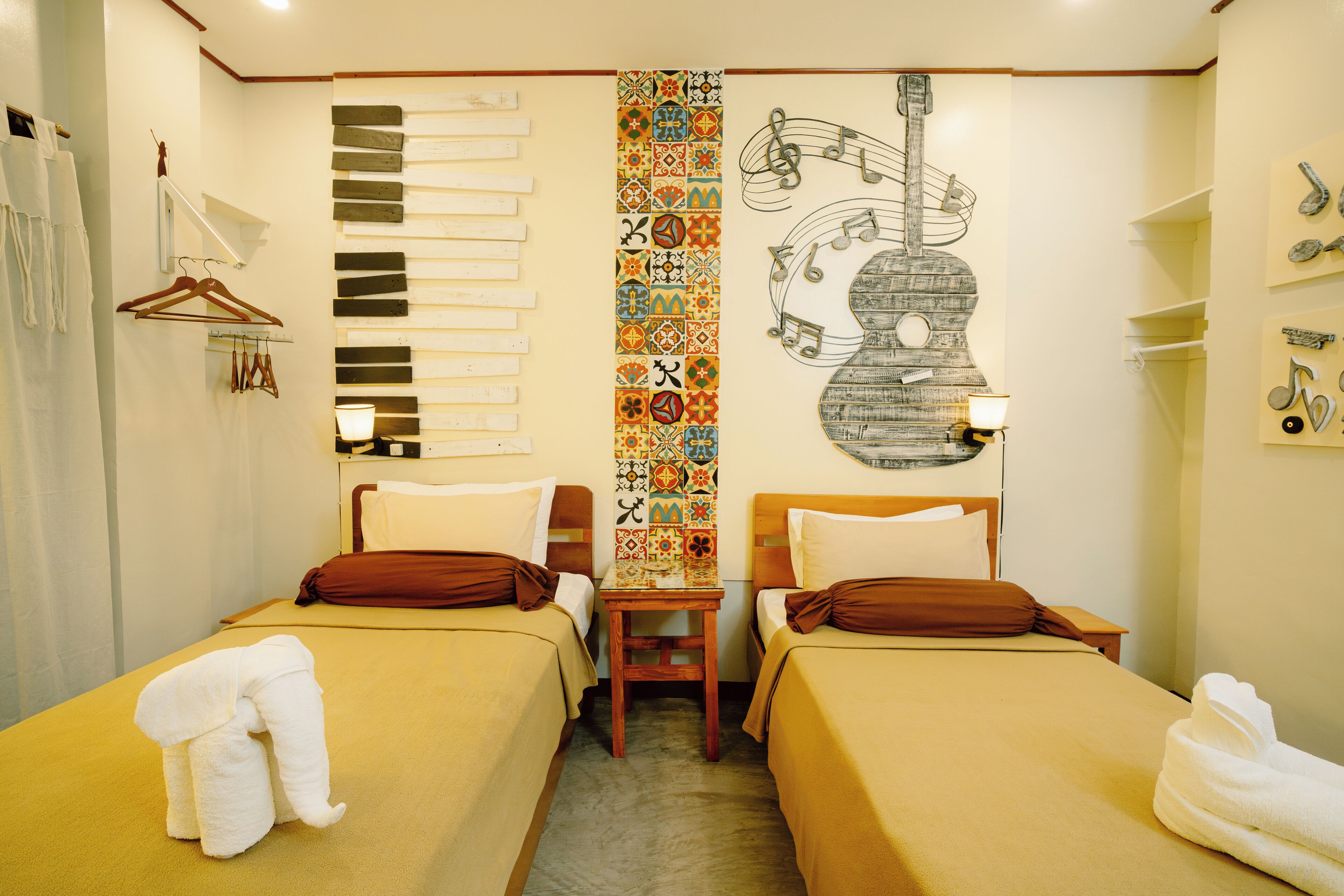 Une chambre d’hôtel avec deux lits, une tête de lit en bois, un mur décoré d’une guitare et de partitions musicales, et un motif vertical aux couleurs vives.