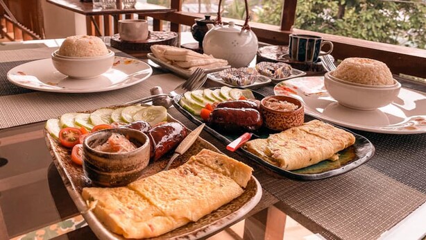 A dining table with various dishes, including rice, flatbreads, and a variety of meats, set on a balcony with a view of trees.