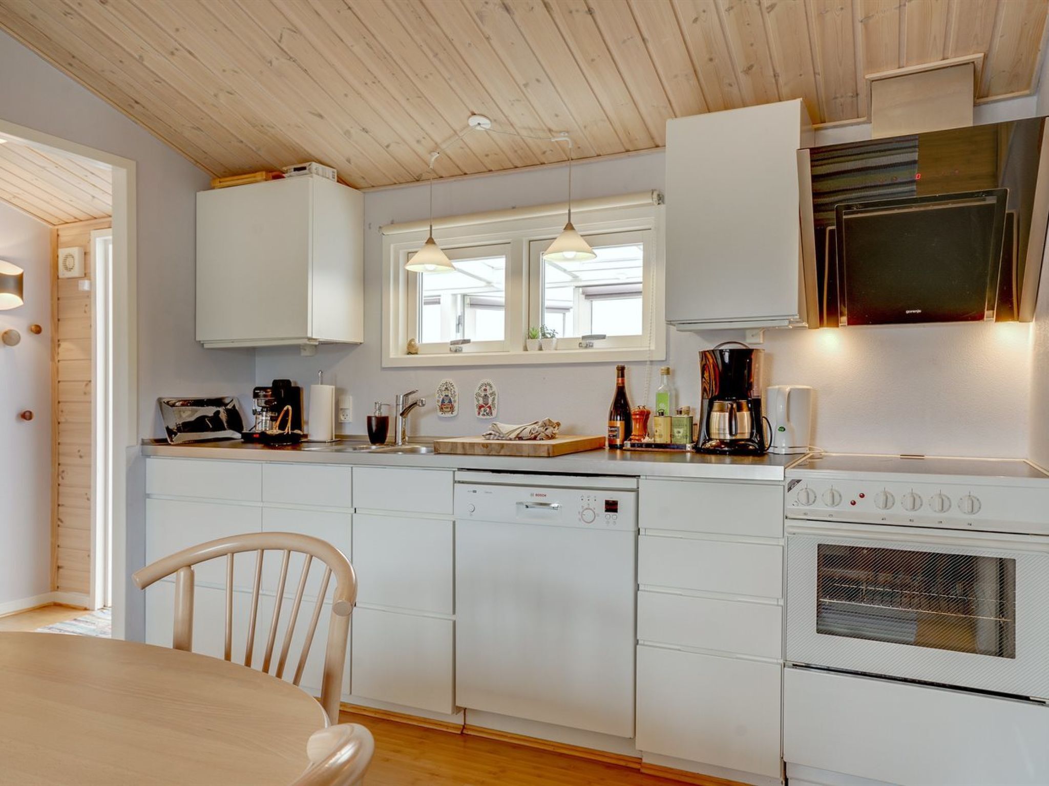 Een moderne keuken met witte kasten, een Bosch vaatwasser, een kookplaat en een magnetron. Er is een houten eettafel met stoelen en hanglampen boven het aanrecht.