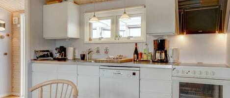 Een moderne keuken met witte kasten, een Bosch vaatwasser, een kookplaat en een magnetron. Er is een houten eettafel met stoelen en hanglampen boven het aanrecht.