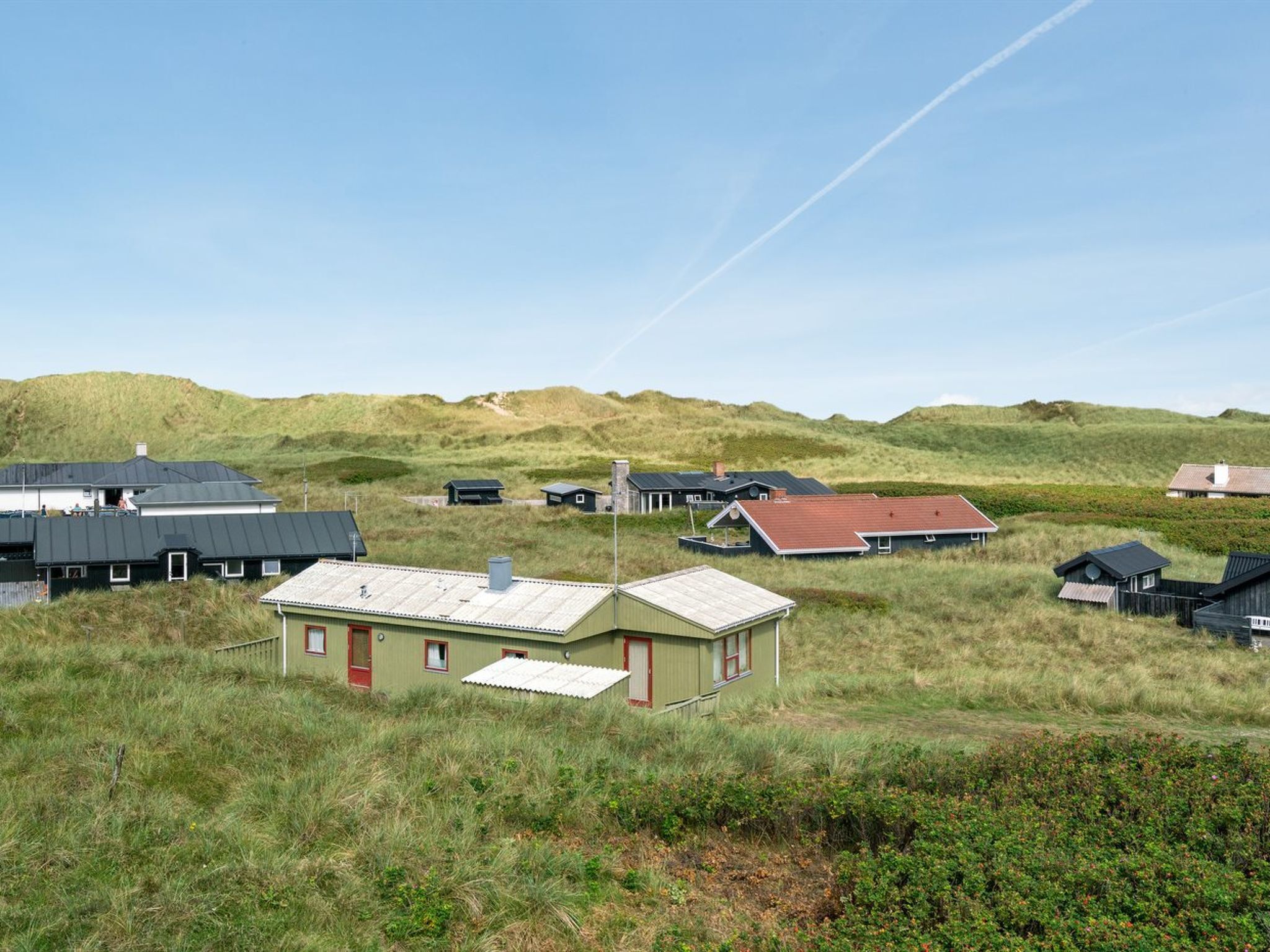 A coastal area with multiple buildings, including a prominent house with a red door and a flat roof, surrounded by grassy dunes and a clear sky.