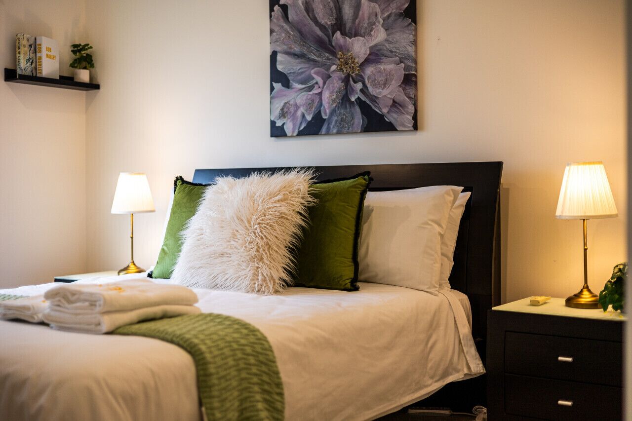 Ein ordentlich bezogenes Bett mit weißen und grünen Kissen, einer flauschigen weißen Decke und einer grünen Wolldecke. Zwei Nachttischlampen, ein Wandbild mit Blumen und ein Regal mit Büchern und einer kleinen Pflanze sind ebenfalls zu sehen.