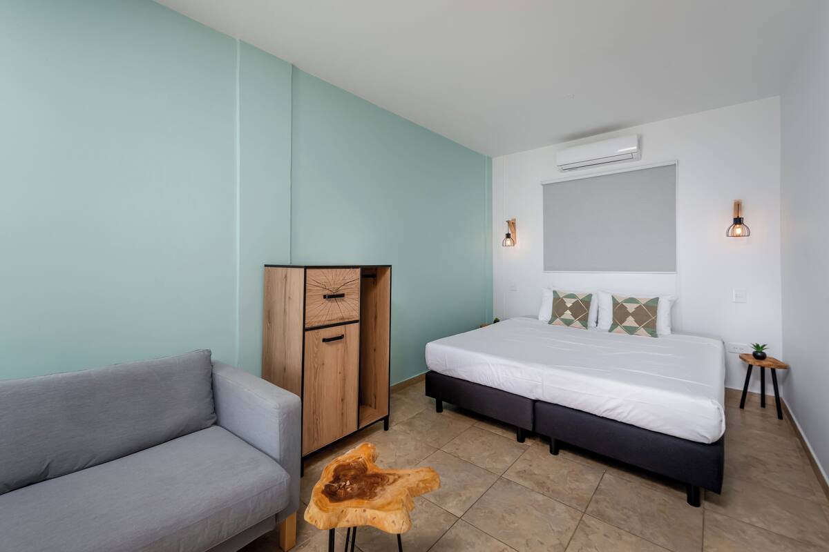 A bedroom with a bed, a grey sofa, a wooden wardrobe, a small stool, and a nightstand with a lamp.
