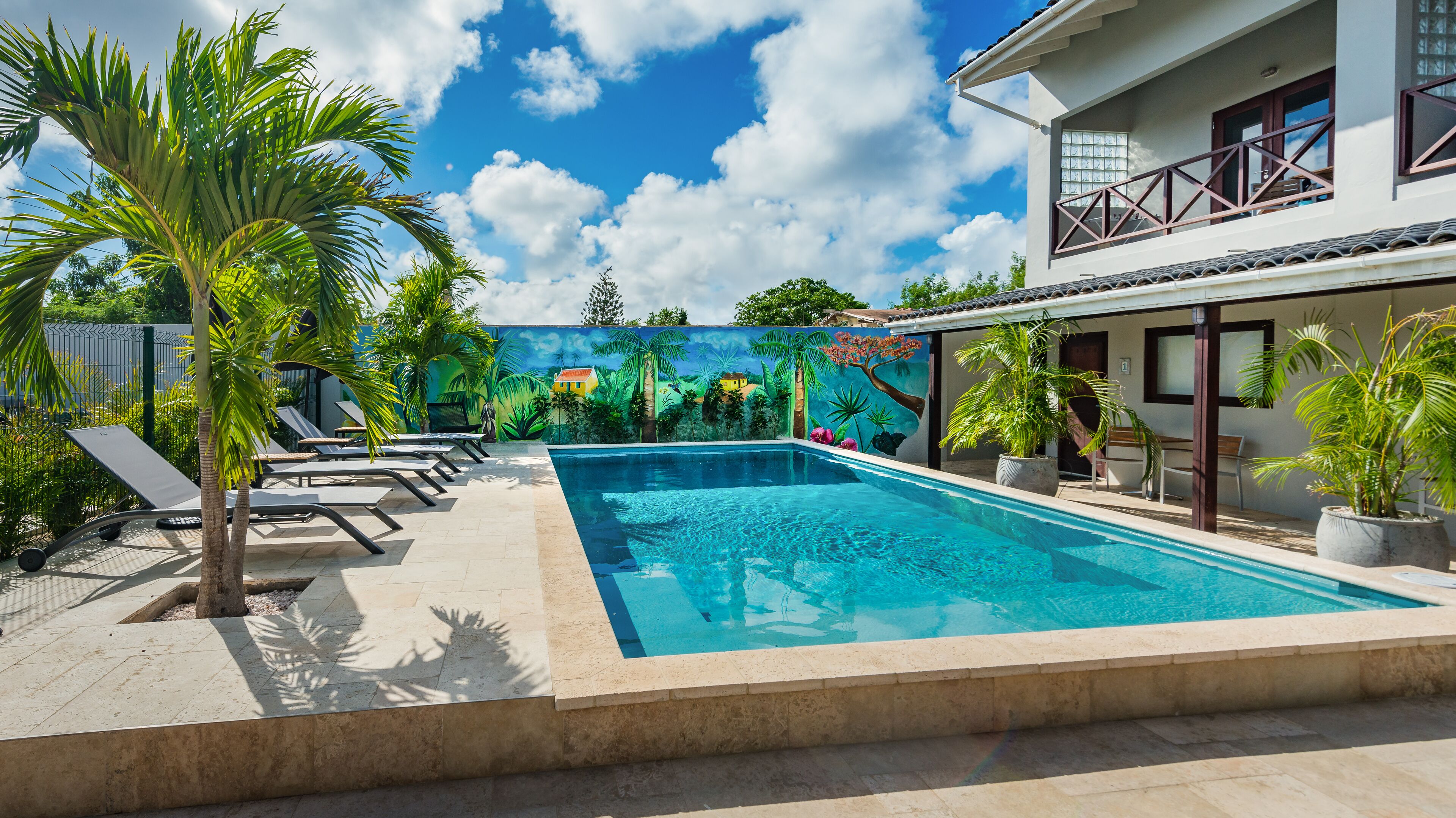 Uma piscina com espreguiçadeiras, palmeiras e um mural com paisagem tropical.