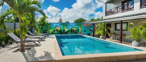 Uma piscina com espreguiçadeiras, palmeiras e um mural com paisagem tropical.