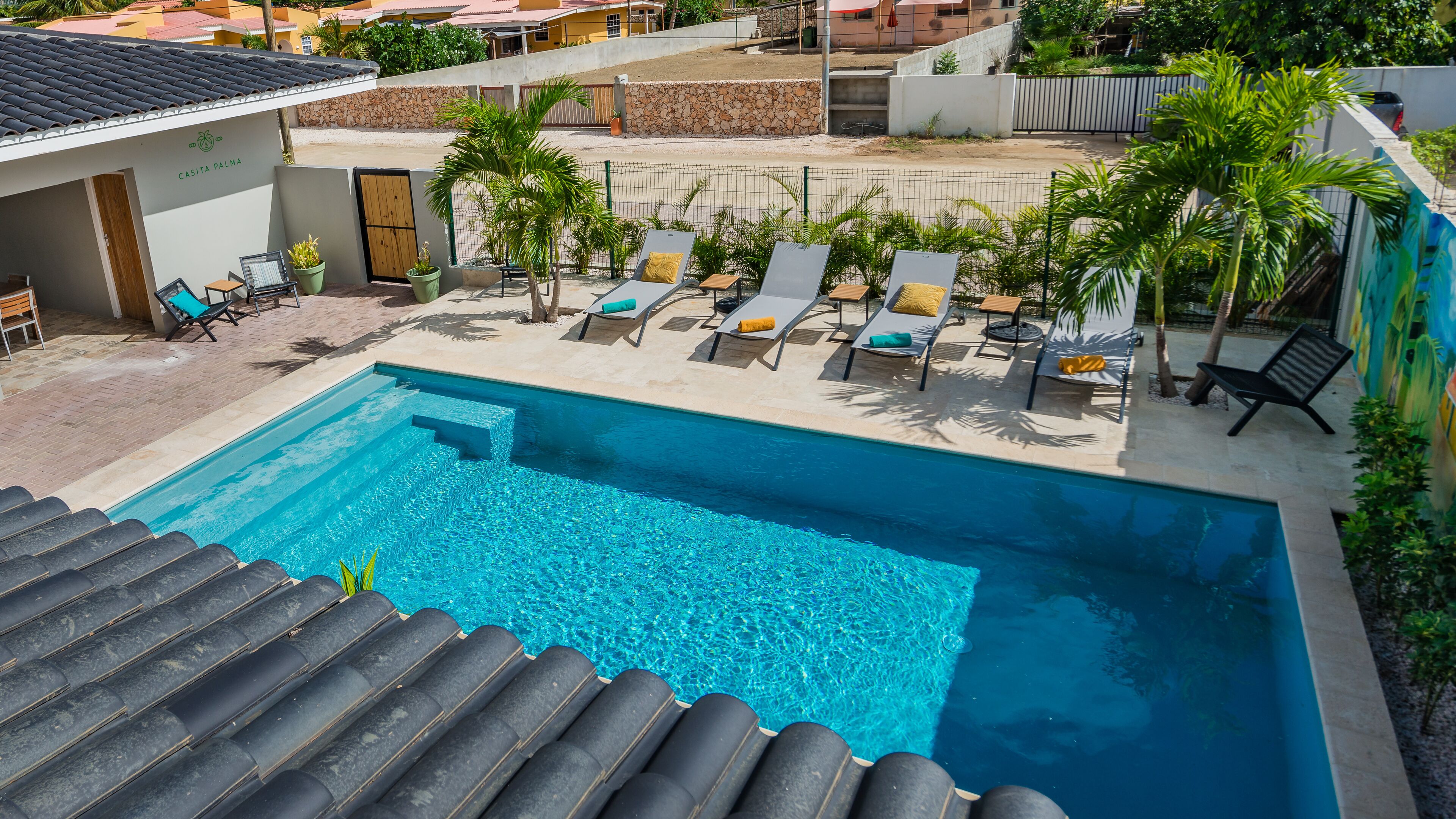Una piscina con sillas de descanso y palmeras, rodeada por un muro de piedra y un edificio con la inscripción 'CASITA PALMA'.
