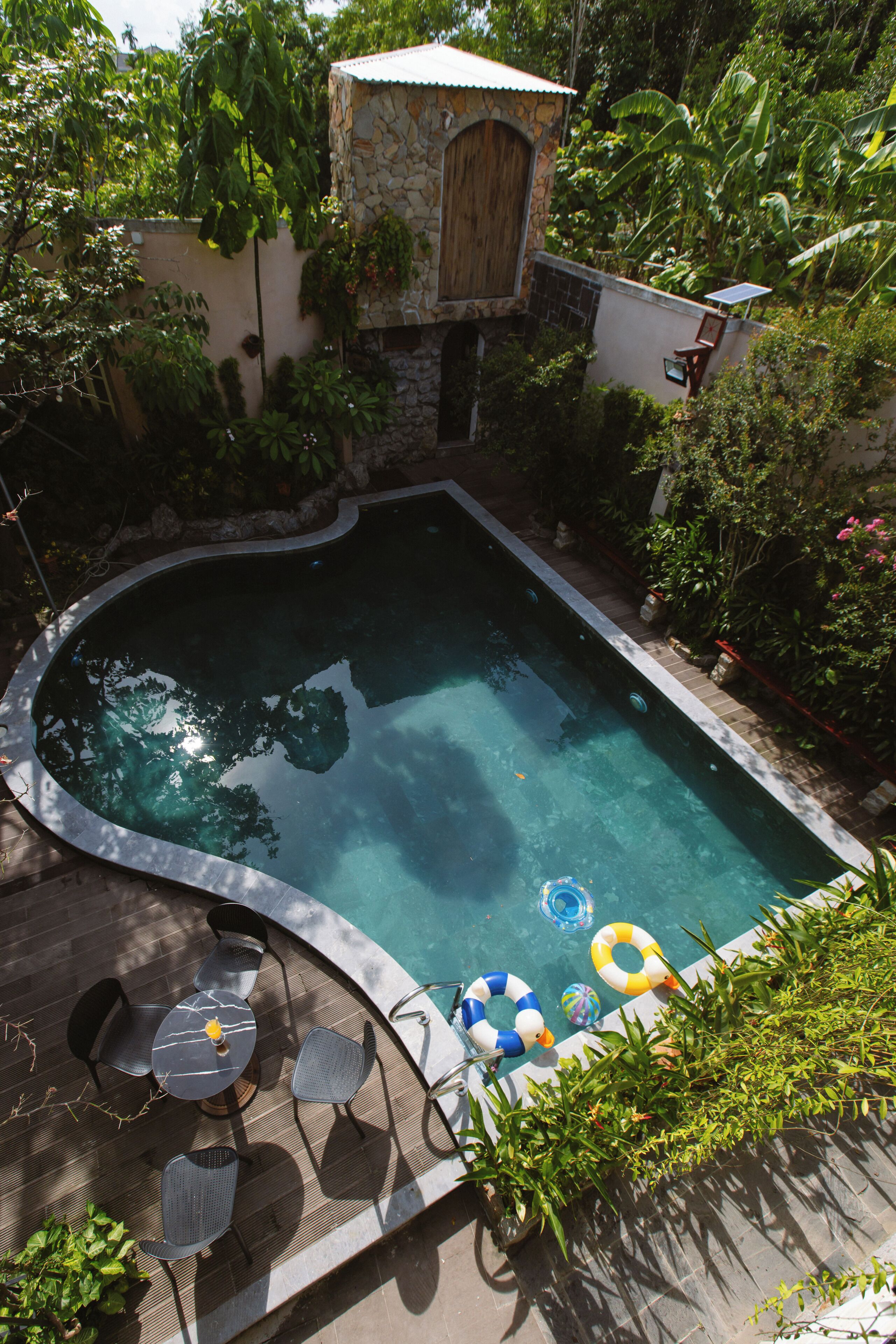 A swimming pool with a stone wall, surrounded by lush greenery and a small stone building.