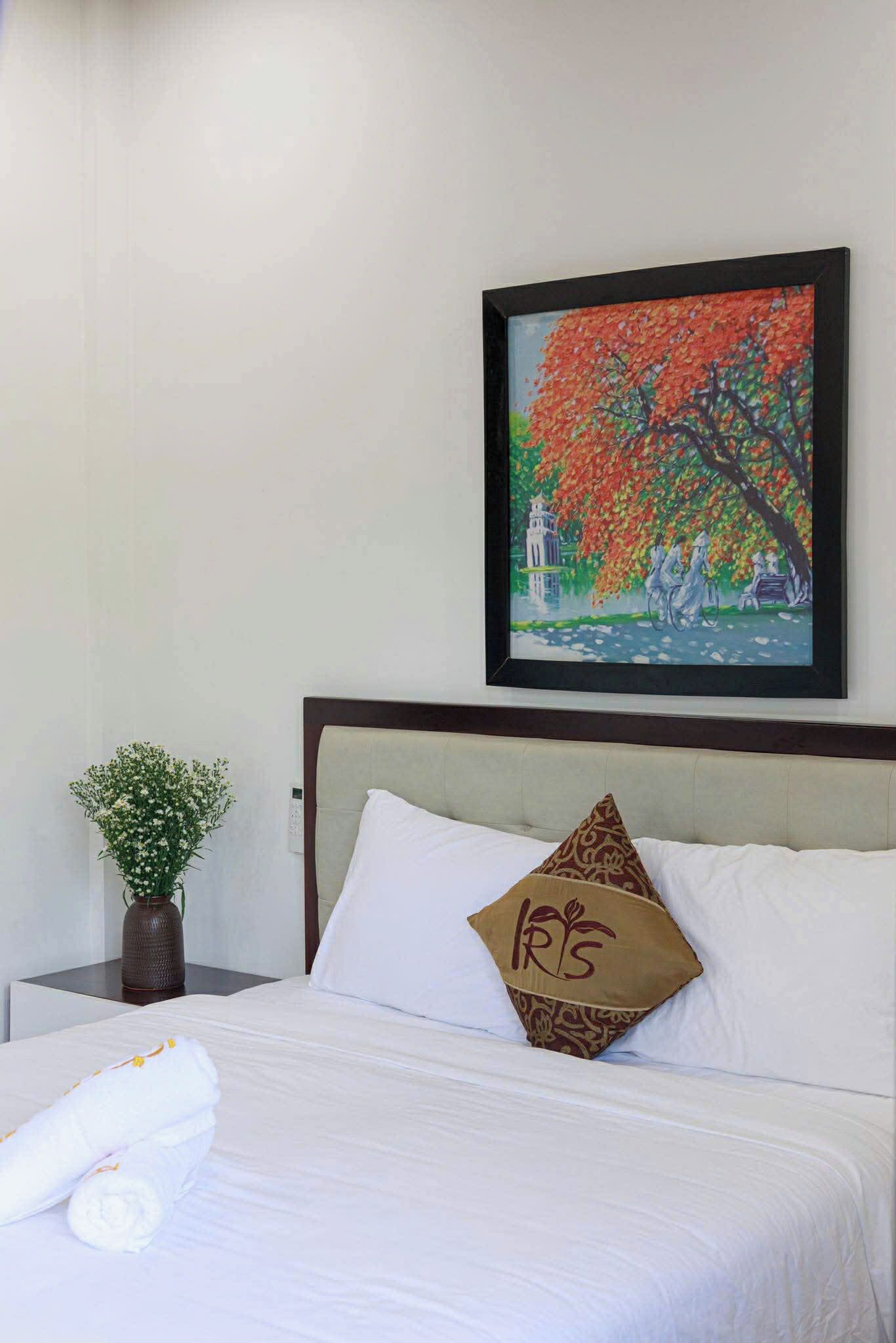 Quarto de hotel com uma cama, um quadro com uma árvore, um vaso com flores e uma almofada estampada.