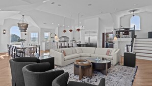 A spacious living area with a large L-shaped sectional sofa, a dining table with chairs, and a kitchen in the background. - 2876 - The Blue Martini - Oceanfront Luxury Retreat w/ Pool & Sauna (Duck)