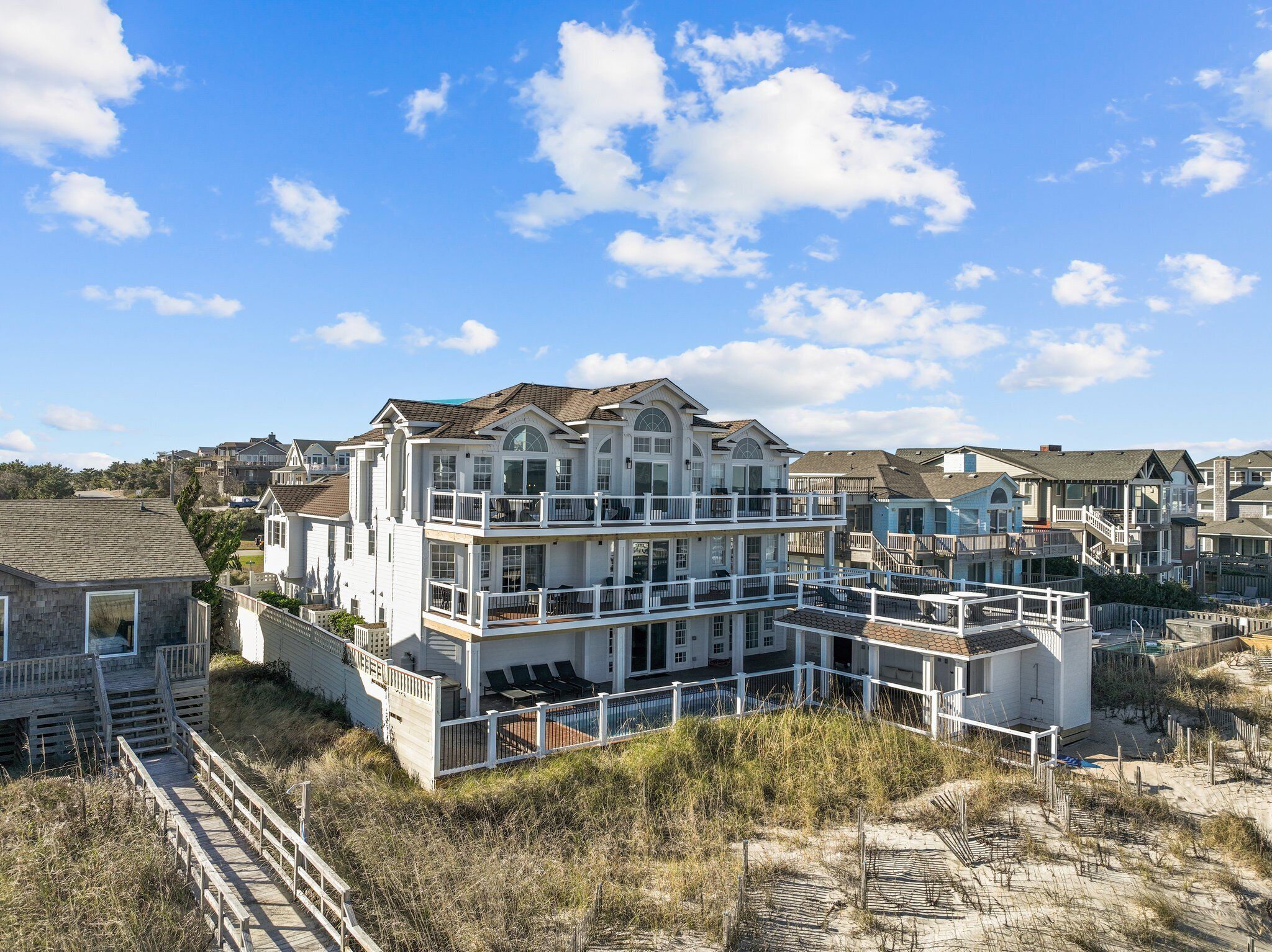 A multi-story beachfront house with balconies, a swimming pool, and a wooden boardwalk leading to the beach.