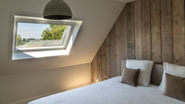 Un dormitorio con techo inclinado, un tragaluz, una cama con ropa de cama blanca y una pared de madera.