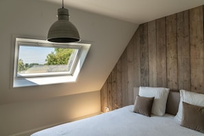 A bedroom with a sloped ceiling, a skylight, a bed with white bedding, and a wooden wall.