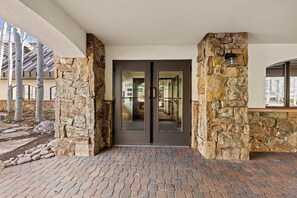 A stone-clad entrance with a glass door, a brick pathway, and birch trees in the background.