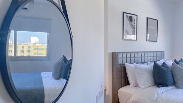 A hotel room with a large round mirror, a bed with white bedding and multiple pillows, two framed pictures on the wall, and a window with a view of buildings. - Phaedrus Living Plateia Suite Grigio (Nicosia)