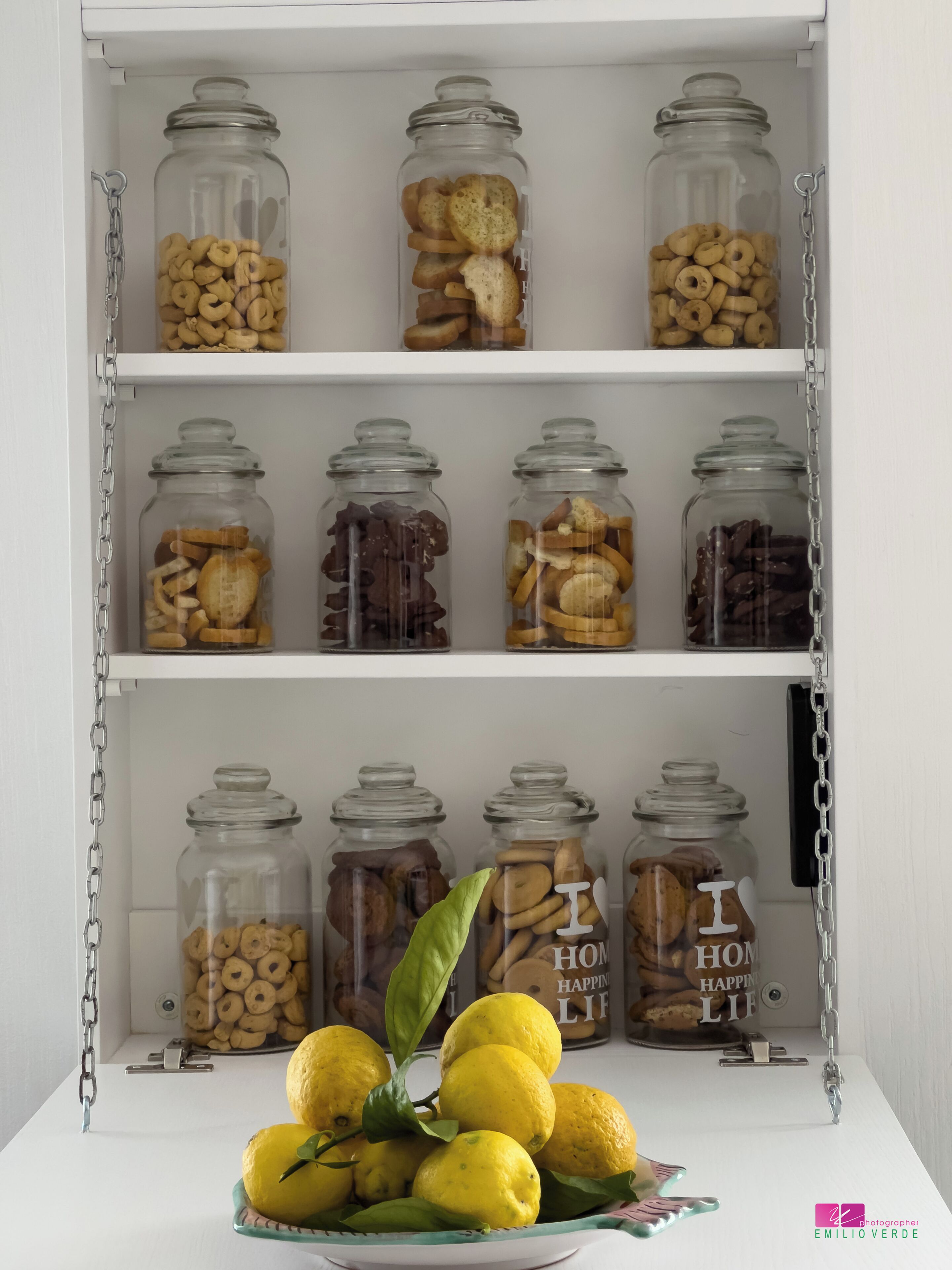 Ein weißes Regal mit mehreren Glaskolben, gefüllt mit verschiedenen Snacks und Keksen, und eine Zitronenschale unten.