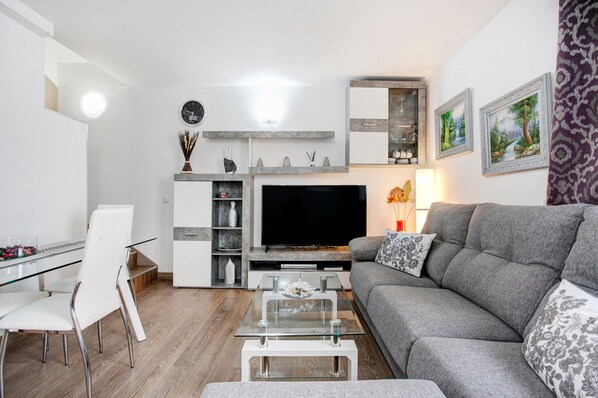 Un salon moderne avec un téléviseur à écran plat, un canapé gris, une table basse en verre, une table à manger blanche et des chaises.