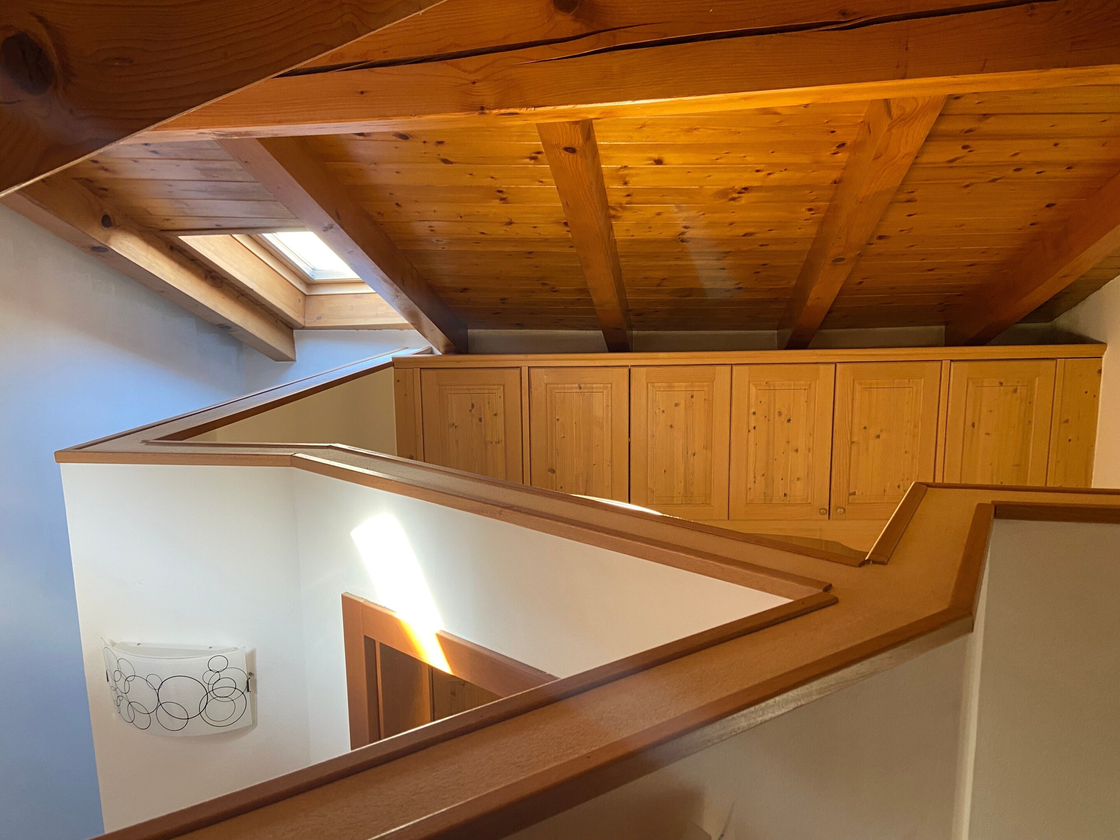 A wooden staircase leading to an attic with a skylight, wooden cabinets, and a white wall with a circular design.