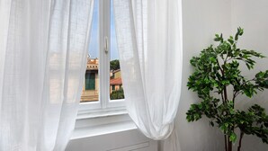 A window with sheer white curtains, a view of buildings outside, and a potted plant on the right. - Rosa Corallo Deluxe – Historic Center Apartment with Wi-Fi and Air Conditioning (Varazze)