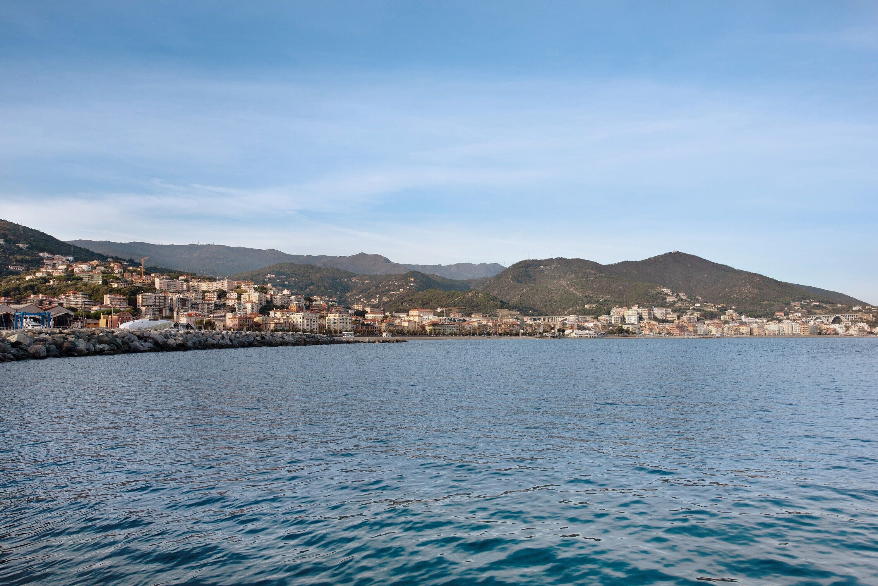 A coastal cityscape with buildings along the waterfront, a clear sky, and mountains in the background.