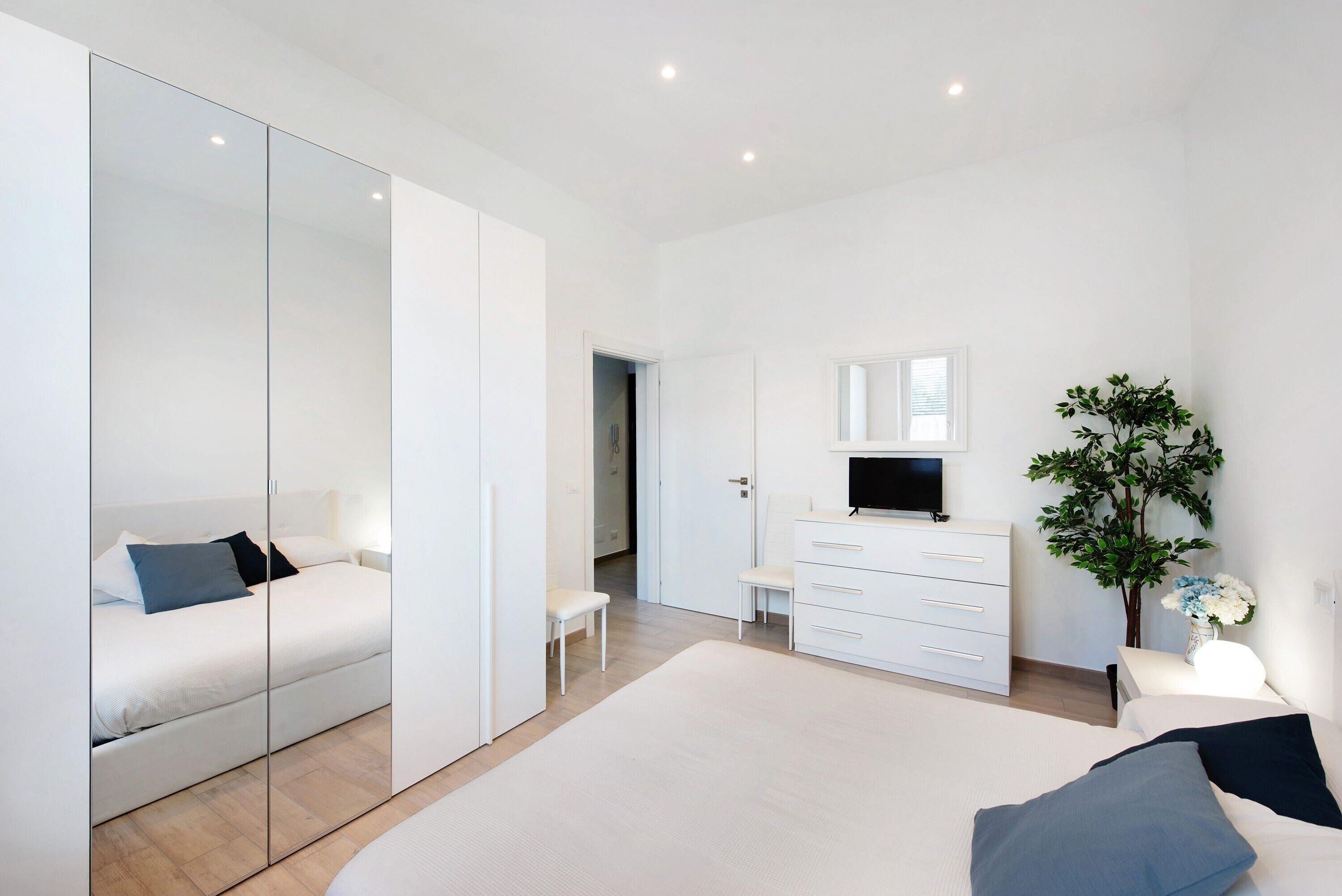 Une chambre moderne avec un grand lit, une armoire avec un miroir, une commode, un banc et une plante en pot.