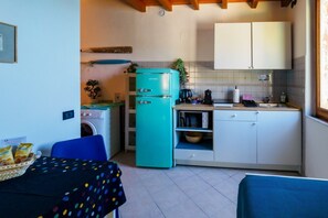 Une cuisine compacte avec un réfrigérateur turquoise, un lave-linge et un four à micro-ondes. On y trouve une table à manger recouverte d’une nappe à pois et une chaise bleue.