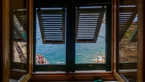 Ein Fenster mit Rollläden, das auf ein Gewässer und eine felsige Küste mit Menschen blickt.