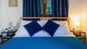 Ein ordentlich bezogenes Bett mit blauer Bettdecke und zwei blauen Kissen, ein Nachttisch mit Lampe und ein kleiner Topfpflanze.