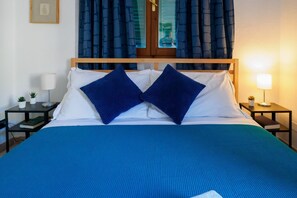 Una cama bien hecha con una colcha azul y dos almohadas azules, una mesita de noche con una lámpara y una pequeña maceta.