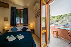 Habitación de hotel con cama, dos lámparas de noche, ventana con cortinas, unidad de aire acondicionado, un cuadro enmarcado y balcón con mesa y silla.
