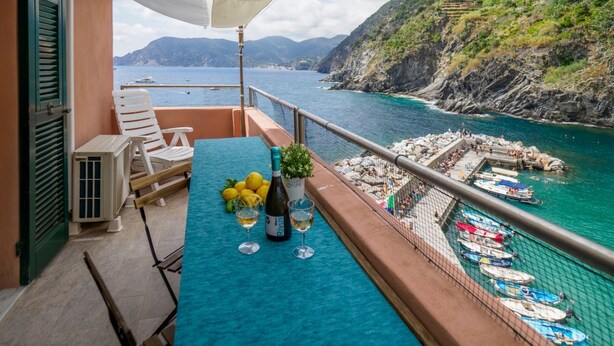 Ein Balkon mit Tisch, gedeckt für Getränke, mit Blick auf eine Küstenlandschaft, Boote und eine felsige Umgebung.