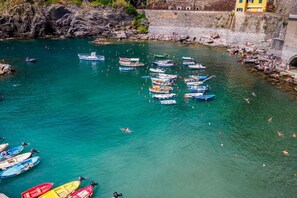 Una zona costera con numerosos barcos amarrados a la orilla, un litoral rocoso y un muro de piedra con un edificio en la parte superior.