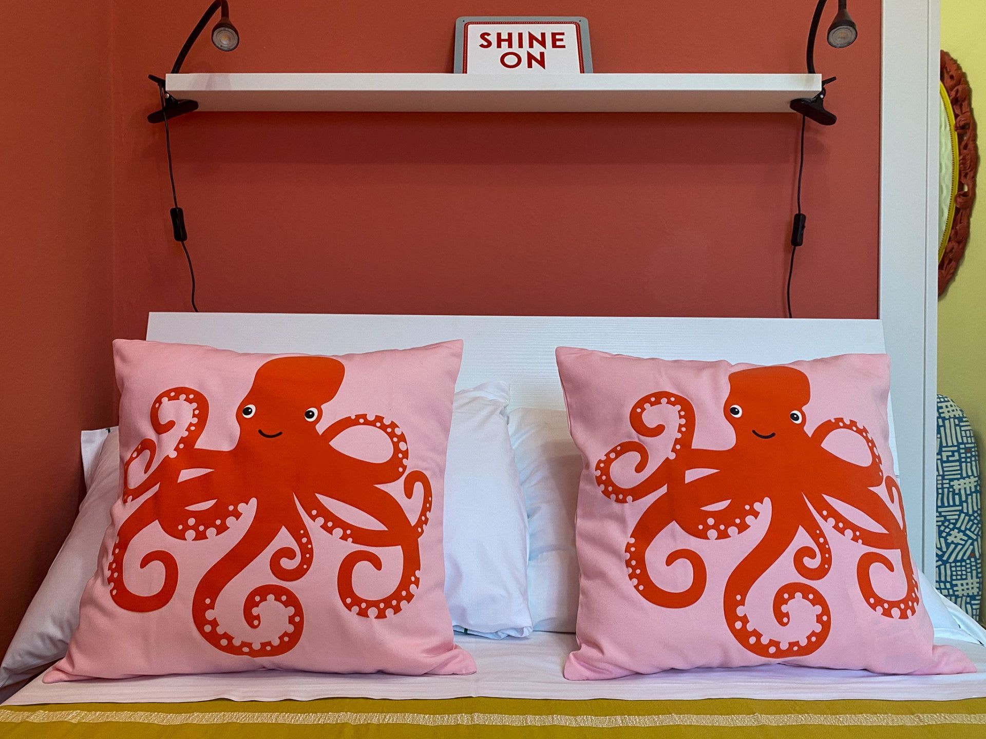 A bed with two pillows featuring an octopus design, a white headboard, and a sign above the headboard that reads 'SHINE ON'.