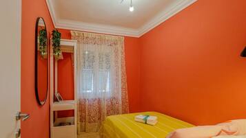 A room with an orange wall, a bed with a yellow bedspread, a white bedside table, a mirror, and a window with patterned curtains.
