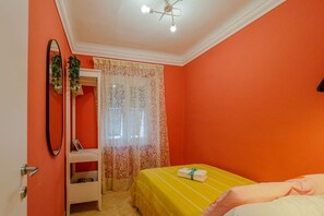 Una habitación con pared naranja, una cama con colcha amarilla, una mesita de noche blanca, un espejo y una ventana con cortinas estampadas.