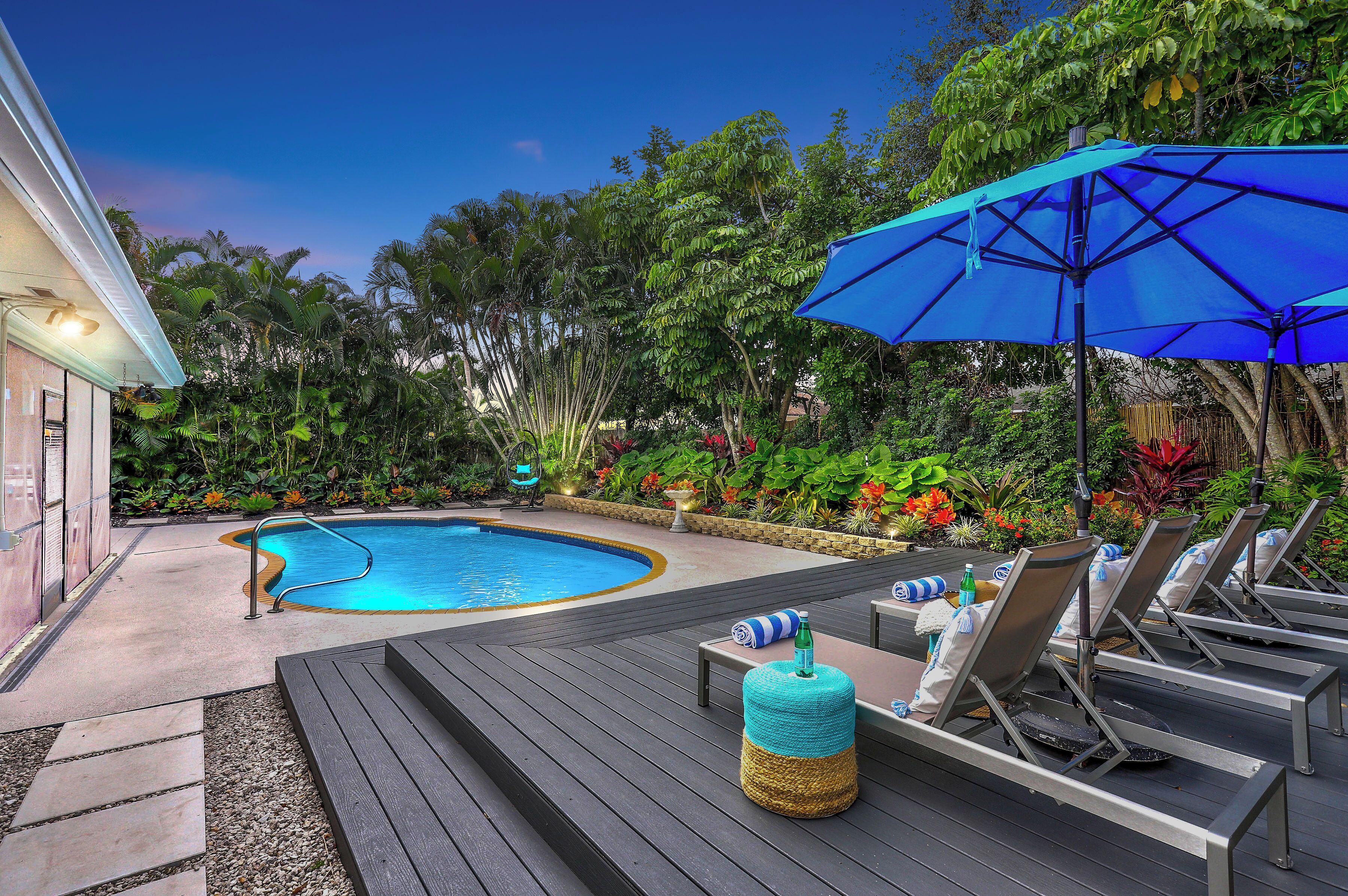 A pool area with lounge chairs and a blue umbrella, surrounded by lush greenery and a wooden deck.