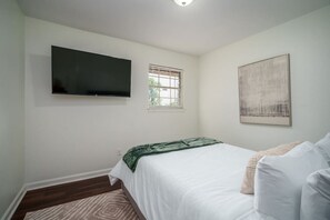 Ein Schlafzimmer mit einem Bett, einem Flachbildfernseher, einem Fenster mit Jalousien und einem Bild an der Wand.