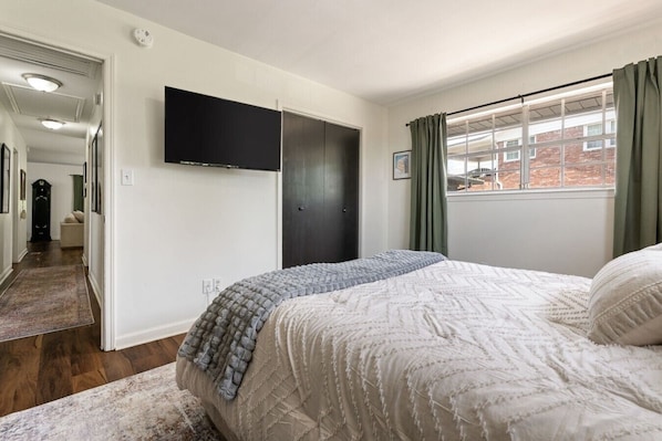 A bedroom with a bed, a flat-screen TV mounted on the wall, a window with curtains, and a hallway leading to another room. - Crestwood Lodge: Hot Tub & Pets! (Birmingham)