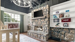 A living room with a stone fireplace, a flat-screen TV mounted on the wall, and a bookshelf filled with books and games.