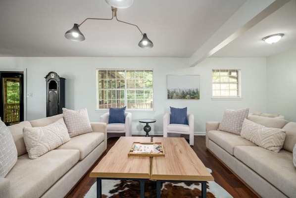 Una sala de estar moderna con dos sofás, una mesa de centro y una pequeña mesa con sillas. Hay ventanas con persianas, una lámpara de techo y una puerta.