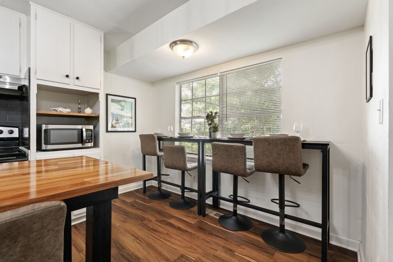 A modern kitchen with a wooden dining table, four bar stools, a microwave, and a window with blinds.