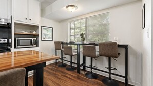 A modern kitchen with a wooden dining table, four bar stools, a microwave, and a window with blinds.