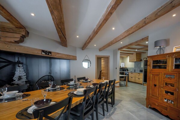 A dining area with a wooden table set for six, surrounded by chairs, with a kitchen in the background.