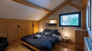 Een slaapkamer met een schuine kap, houten balken, een groot bed met donkere beddengoed, een nachtkastje met een lamp en een raam met uitzicht.