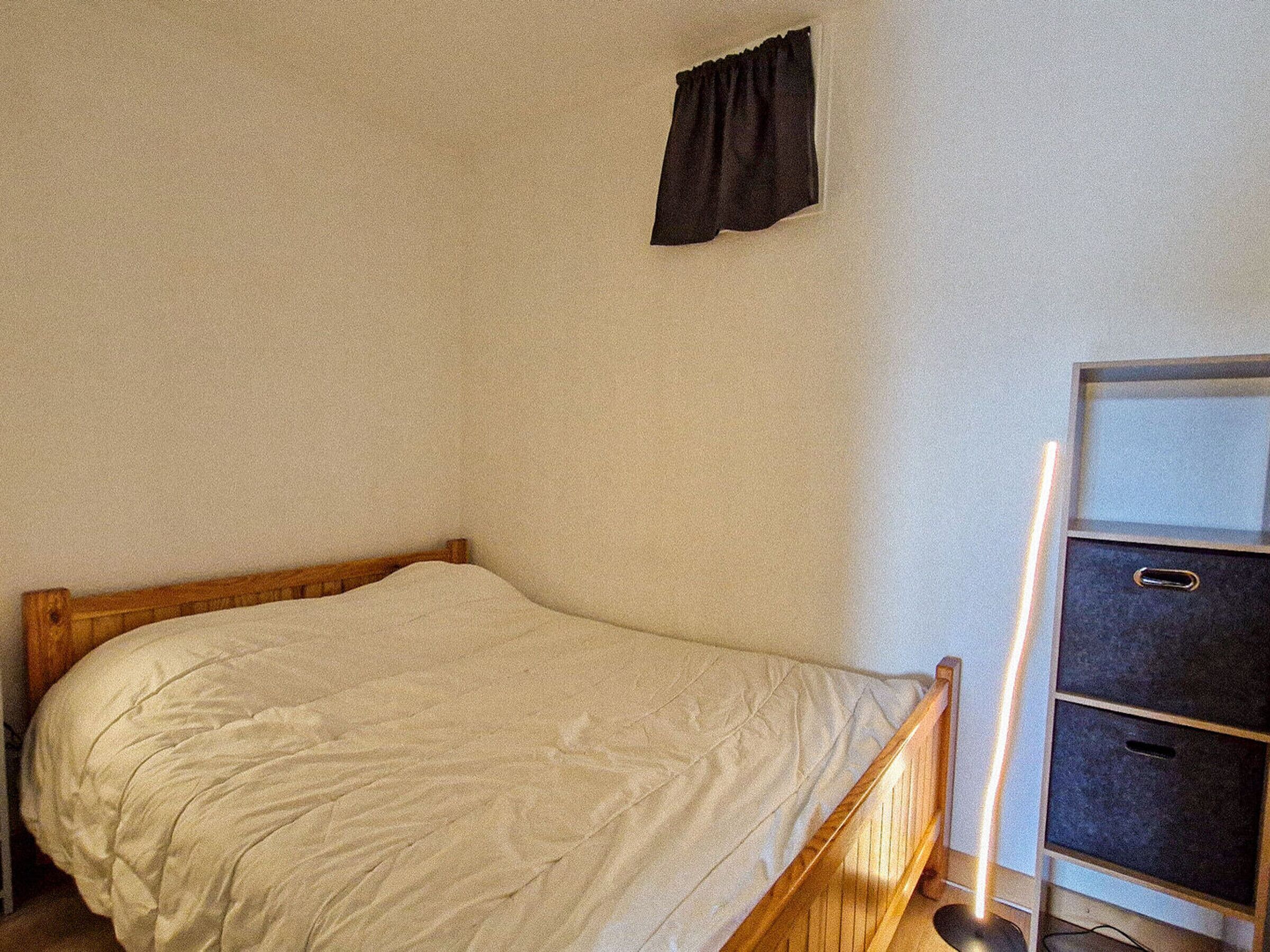 A single bed with a wooden frame and a white mattress in a room with a white wall and a black curtain.