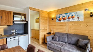 A compact living space with a kitchenette, a gray sofa, wooden walls, and a painting of construction workers. - Renovated studio with cabin in Flaine, sunny balcony (Flaine)