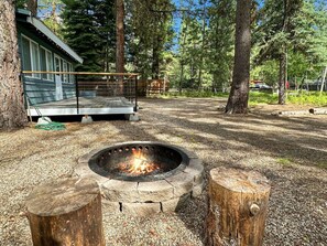 A fire pit with a stone fire ring, surrounded by wooden posts, in a wooded area with a cabin in the background.