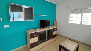 A room with teal walls, a TV on a cabinet, a window with blinds, and a small table. - ACV - Torremar-2nd floor line 5 Front 4 (OROPESA)