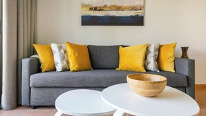 A grey sofa with yellow and white cushions, a wooden bowl on a white table, and a painting on the wall.